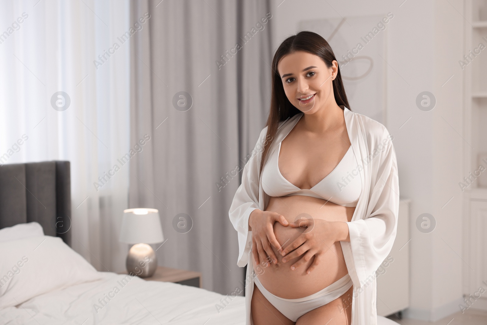 Photo of Beautiful pregnant woman in stylish comfortable underwear and robe making heart with hands on her belly in bedroom, space for text