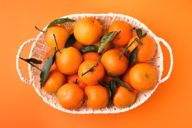 Photo of Fresh ripe tangerines and leaves in basket on orange table, top view