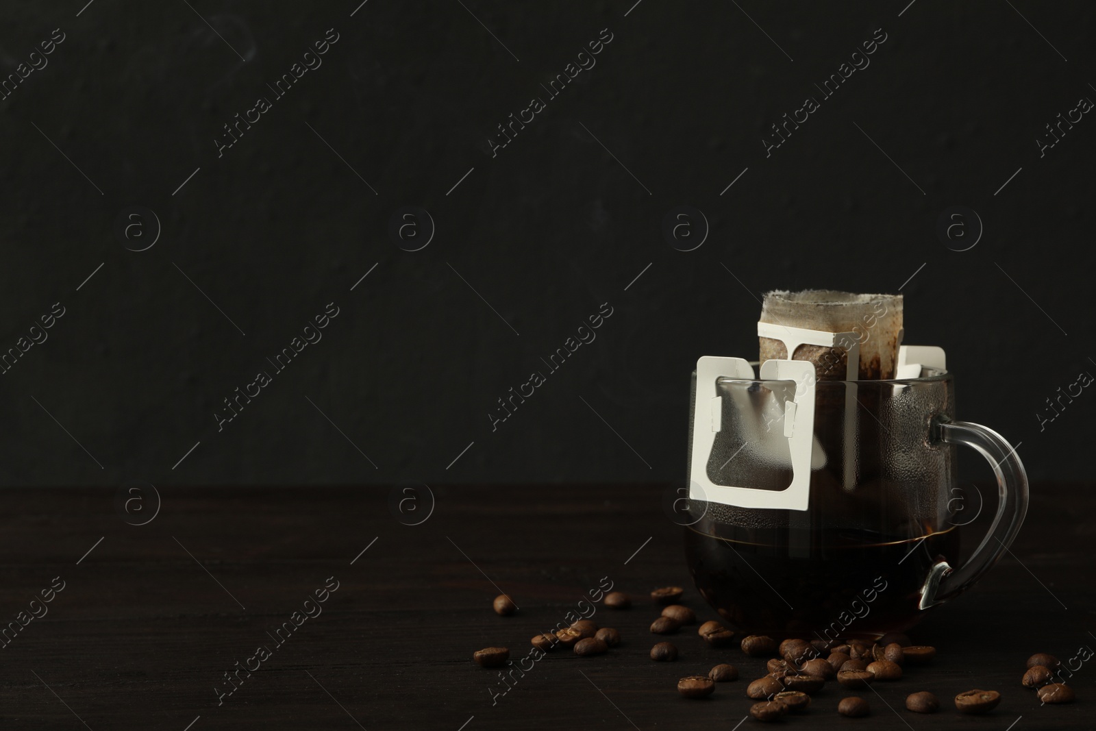 Photo of Glass cup with drip coffee bag and beans on black wooden table. Space for text