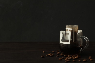 Photo of Glass cup with drip coffee bag and beans on black wooden table. Space for text
