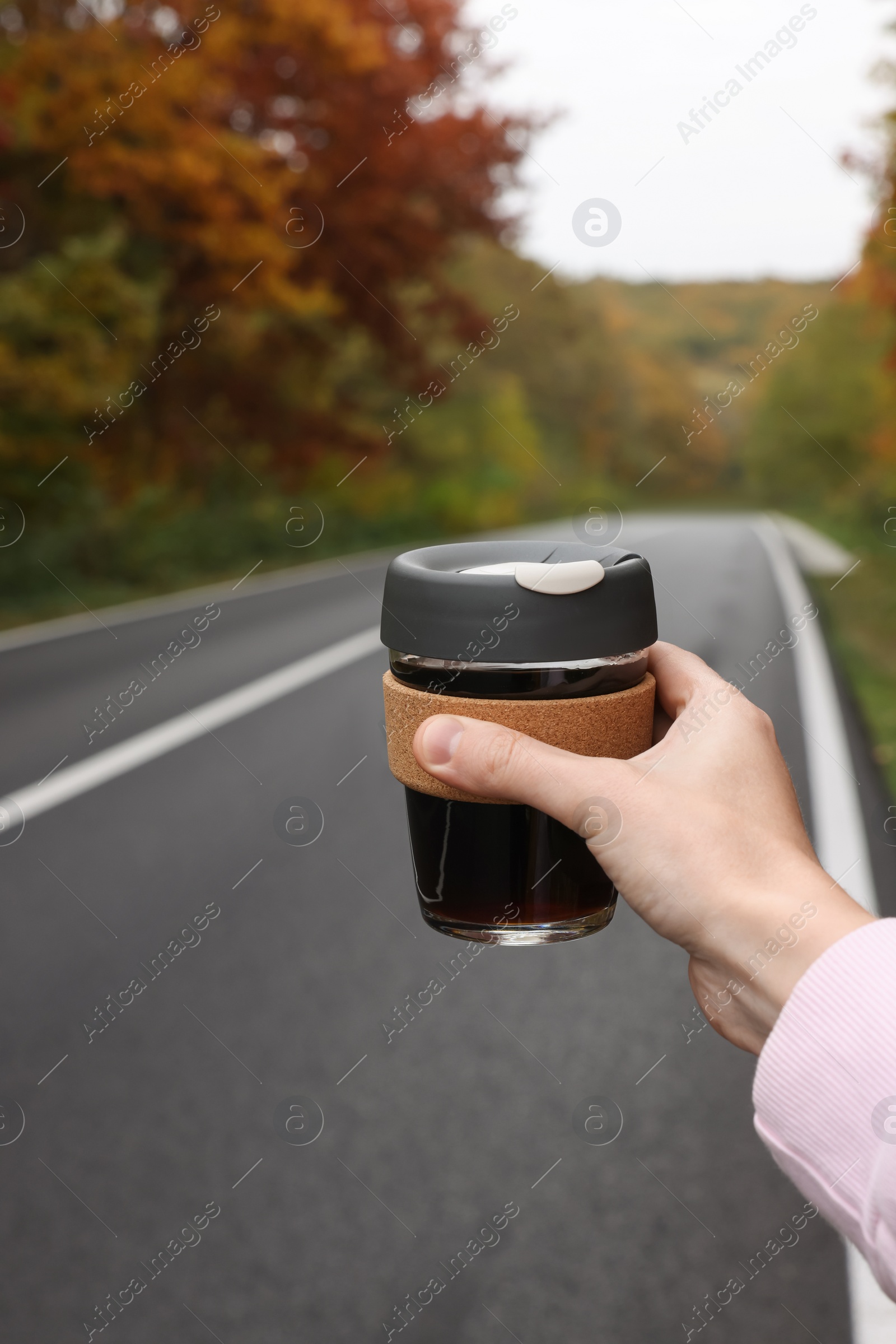 Photo of Woman holding reusable glass cup with hot drink near road outdoors, closeup. Autumn season