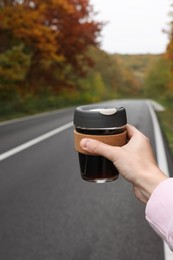 Woman holding reusable glass cup with hot drink near road outdoors, closeup. Autumn season