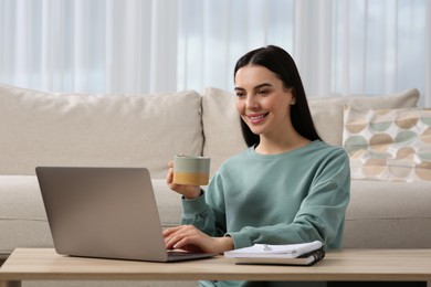 Photo of Happy woman with cup of drink working on laptop at wooden desk in living room