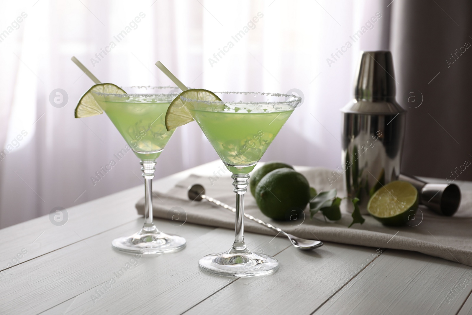 Photo of Delicious Margarita cocktail in glasses, limes and bartender equipment on white wooden table