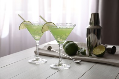 Delicious Margarita cocktail in glasses, limes and bartender equipment on white wooden table