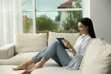 Young woman reading book on sofa at home, space for text