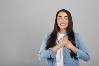 Beautiful grateful woman with hands on chest against light grey background. Space for text