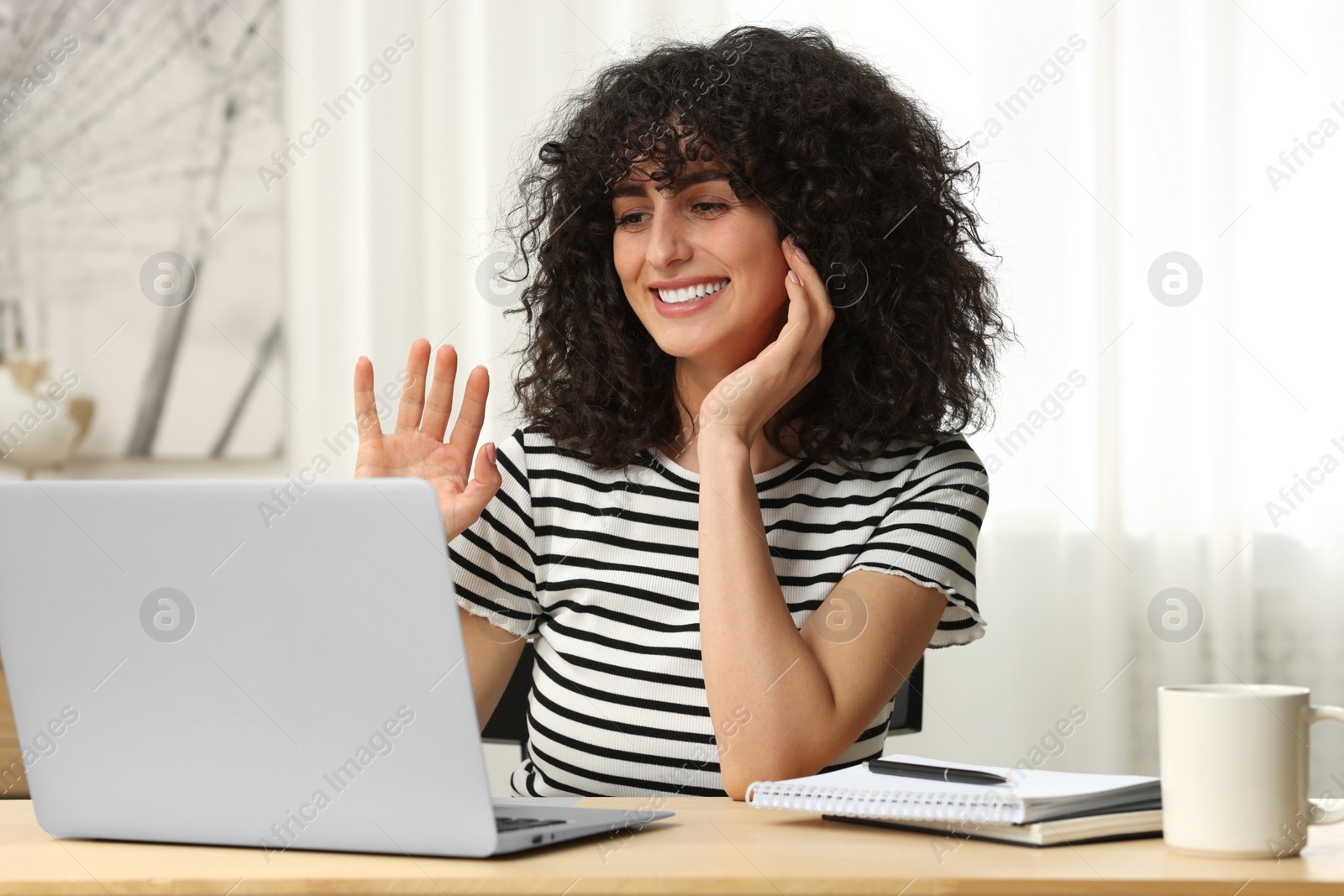 Photo of Happy woman waving hello during video call at table in room