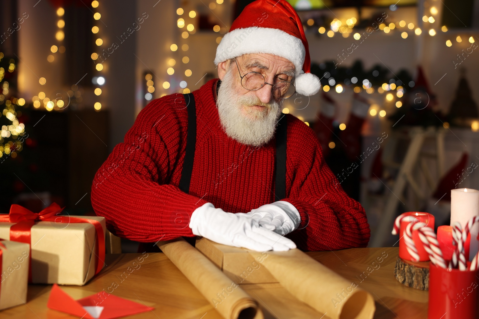 Photo of Santa Claus wrapping gift at his workplace in room decorated for Christmas