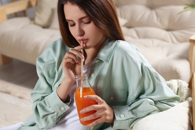 Photo of Beautiful young woman drinking juice from glass bottle on sofa at home