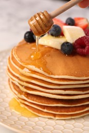 Pouring honey onto delicious pancakes with fresh berries and butter on plate, closeup