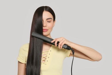 Beautiful woman using hair iron on light grey background