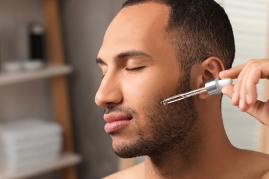 Handsome man applying cosmetic serum onto face in bathroom, closeup