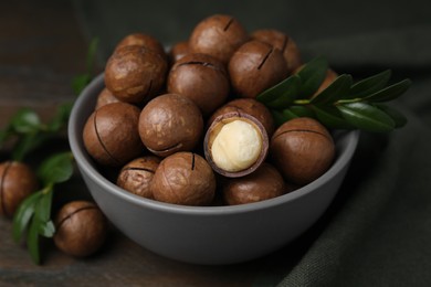 Photo of Tasty Macadamia nuts in bowl and green twigs on table, closeup