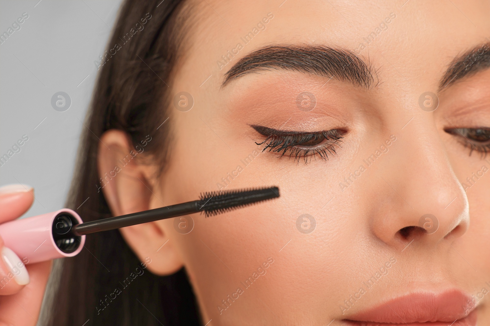 Photo of Beautiful young woman applying mascara on grey background, closeup
