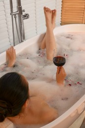 Photo of Woman with glass of wine taking bath in tub with foam and rose petals indoors, back view
