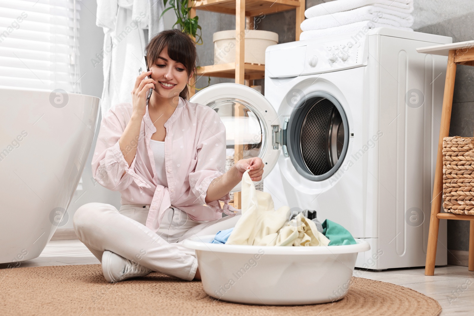 Photo of Happy young housewife with laundry talking on smartphone near washing machine at home