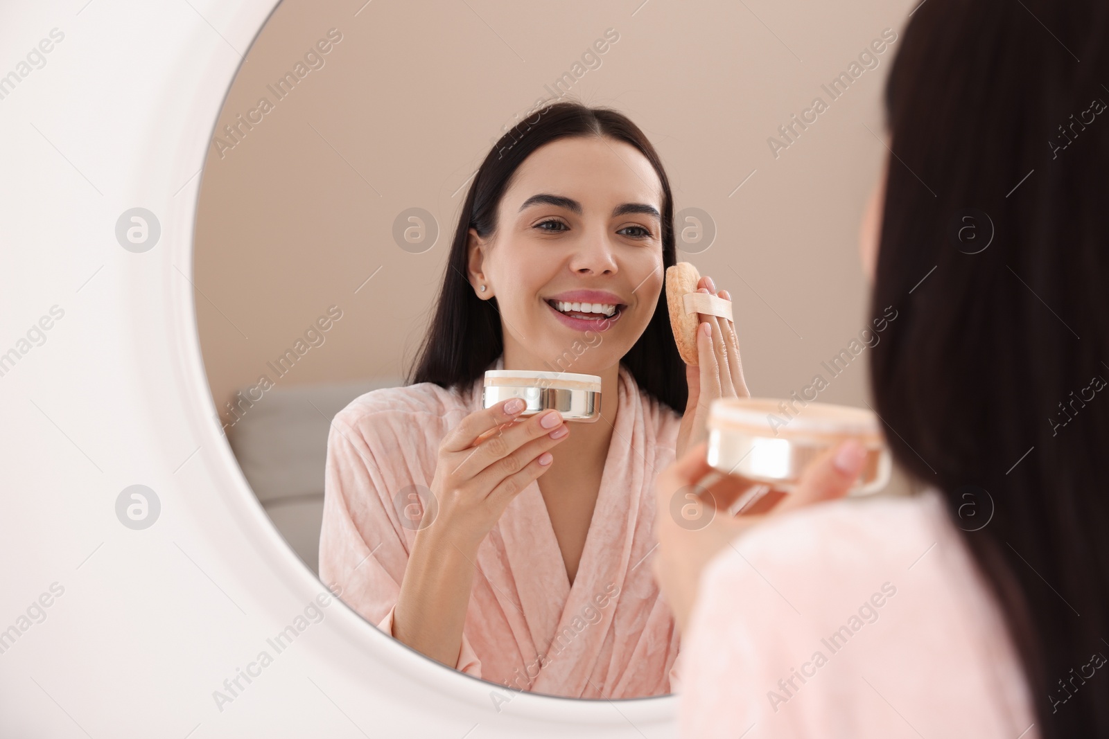 Photo of Beautiful young woman applying powder with puff applicator near mirror indoors