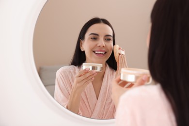 Beautiful young woman applying powder with puff applicator near mirror indoors