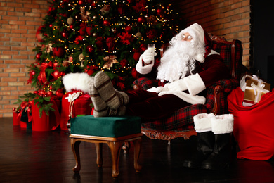 Photo of Santa Claus with glass of milk resting in armchair near Christmas tree