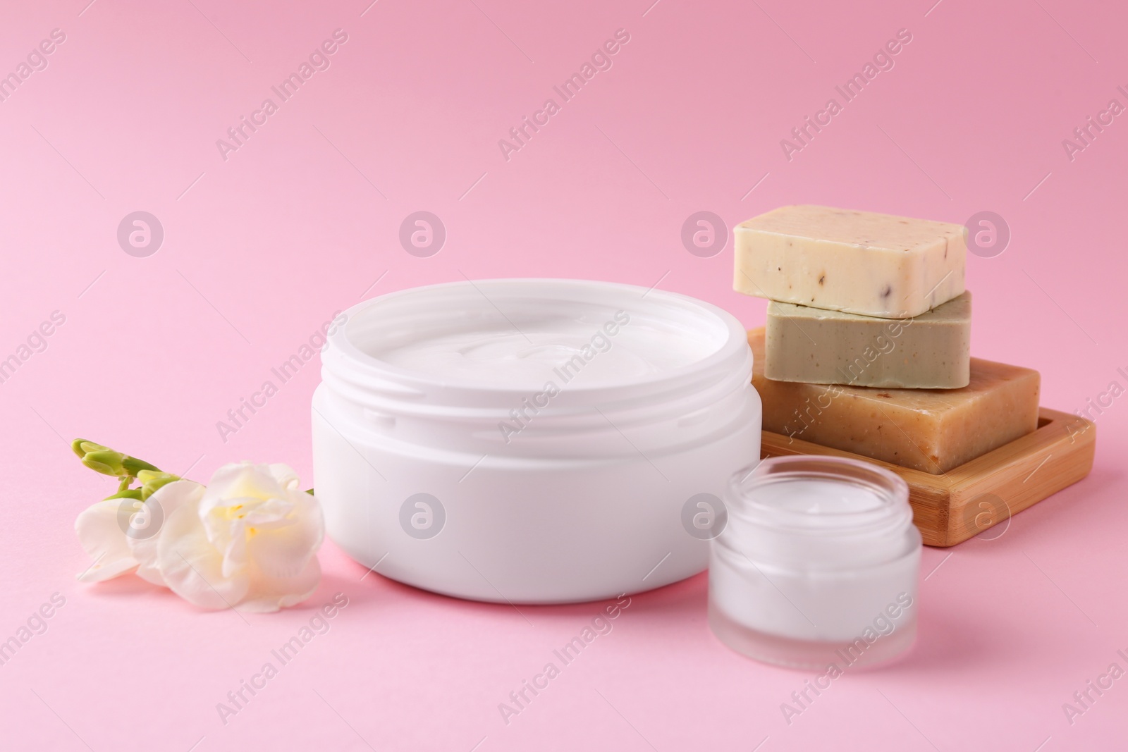 Photo of Moisturizing cream in open jars, soap bars and freesia flower on pink background. Body care products