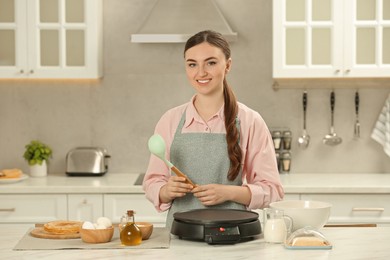 Photo of Happy woman with ladle and electric crepe maker at white marble table in kitchen
