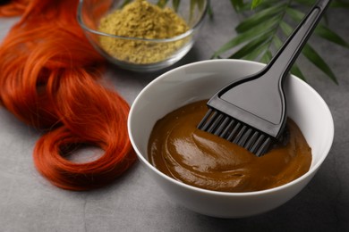 Photo of Bowl of henna cream, brush and red strand on light grey table, closeup. Natural hair coloring