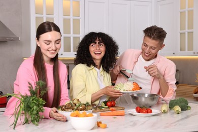 Photo of Friends cooking healthy vegetarian meal at white marble table in kitchen