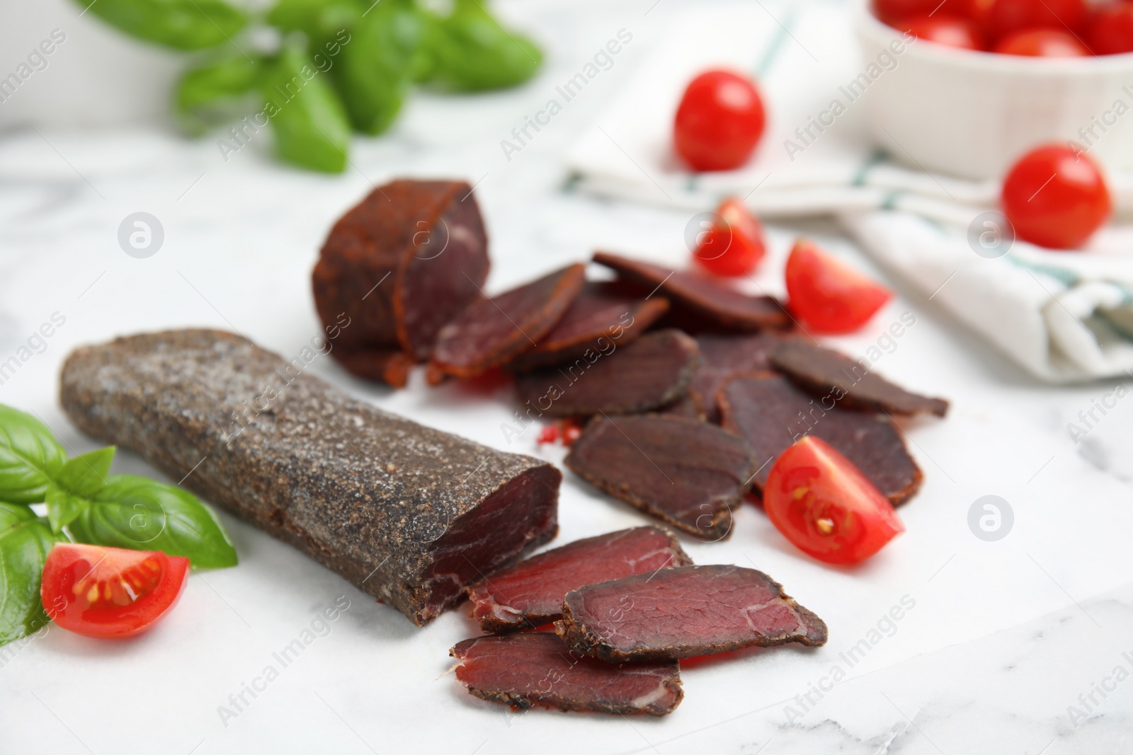 Photo of Delicious dry-cured beef basturma with basil and tomatoes on white marble table, closeup