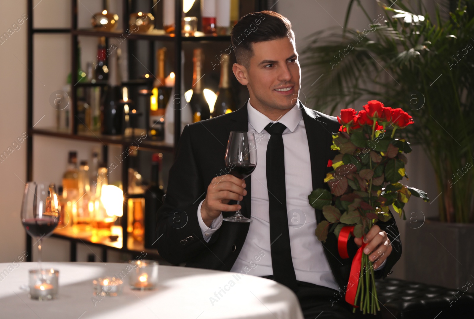 Photo of Happy man with roses and glass of wine waiting for his girlfriend in restaurant on Valentine's day