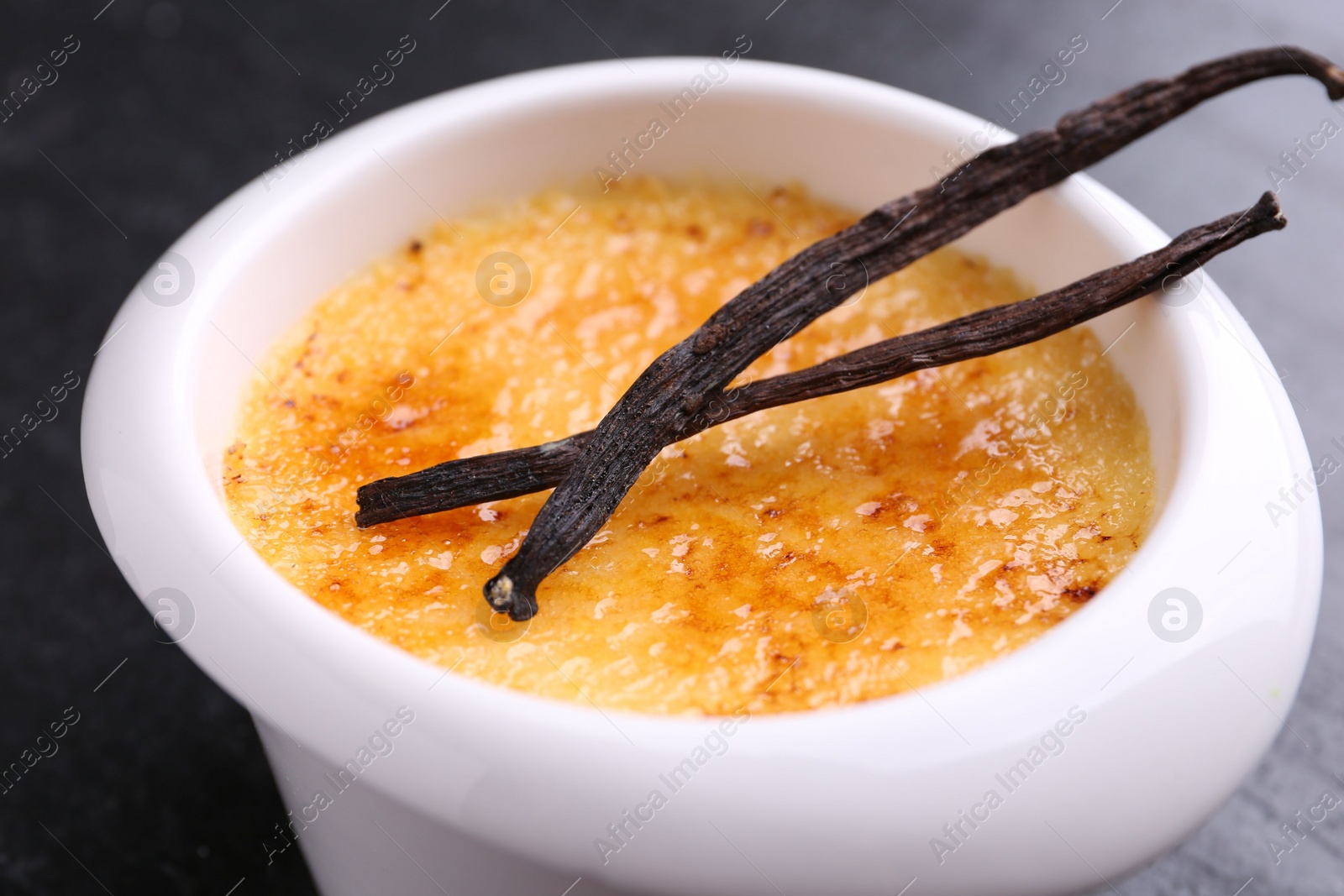 Photo of Delicious creme brulee in bowl and vanilla pods on dark gray table, closeup