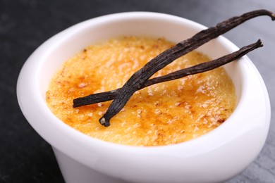Photo of Delicious creme brulee in bowl and vanilla pods on dark gray table, closeup
