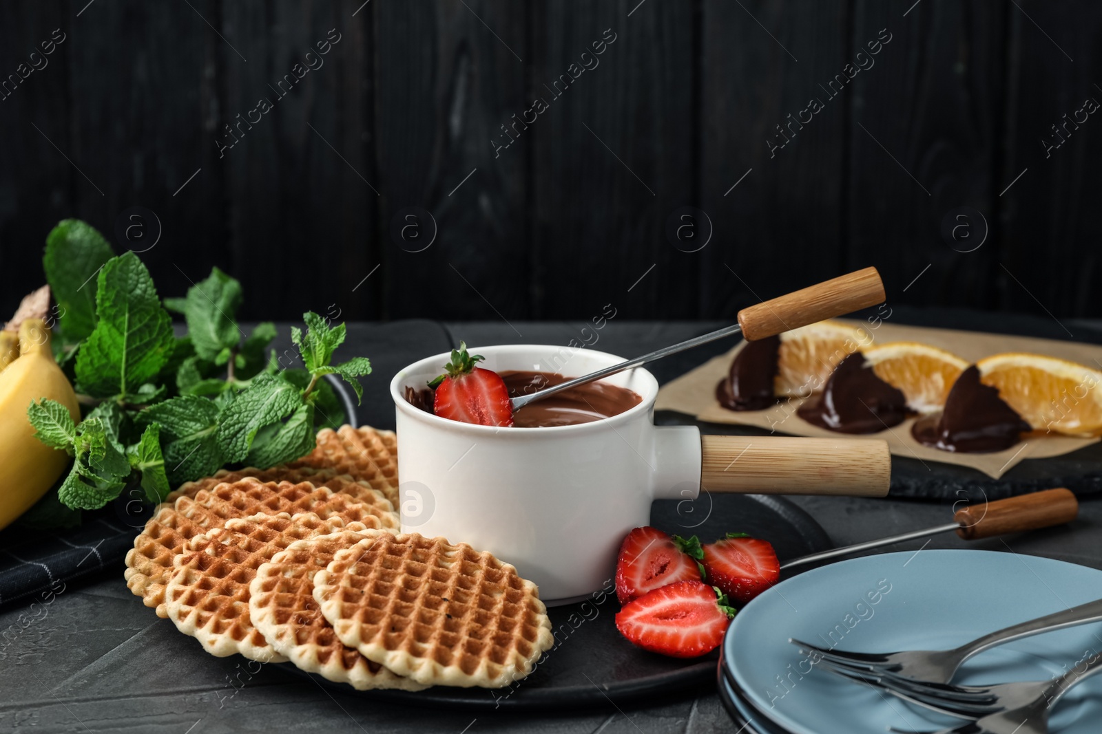 Photo of Dipping strawberry into fondue pot with milk chocolate on grey table against black wooden background