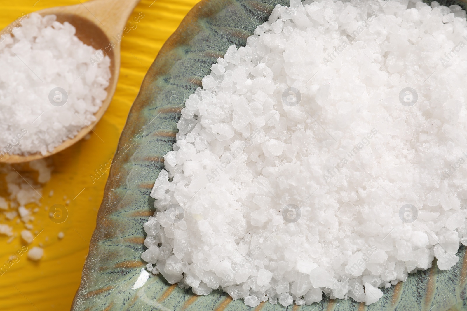 Photo of Natural sea salt in bowl and spoon on yellow wooden table, closeup
