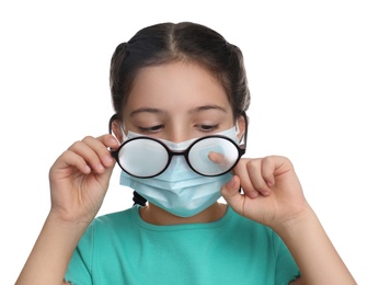 Little girl wiping foggy glasses caused by wearing medical face mask on white background. Protective measure during coronavirus pandemic