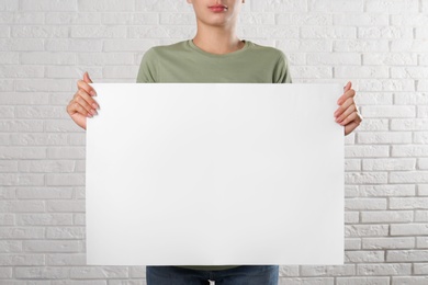 Woman holding blank poster near white brick wall, closeup. Mockup for design