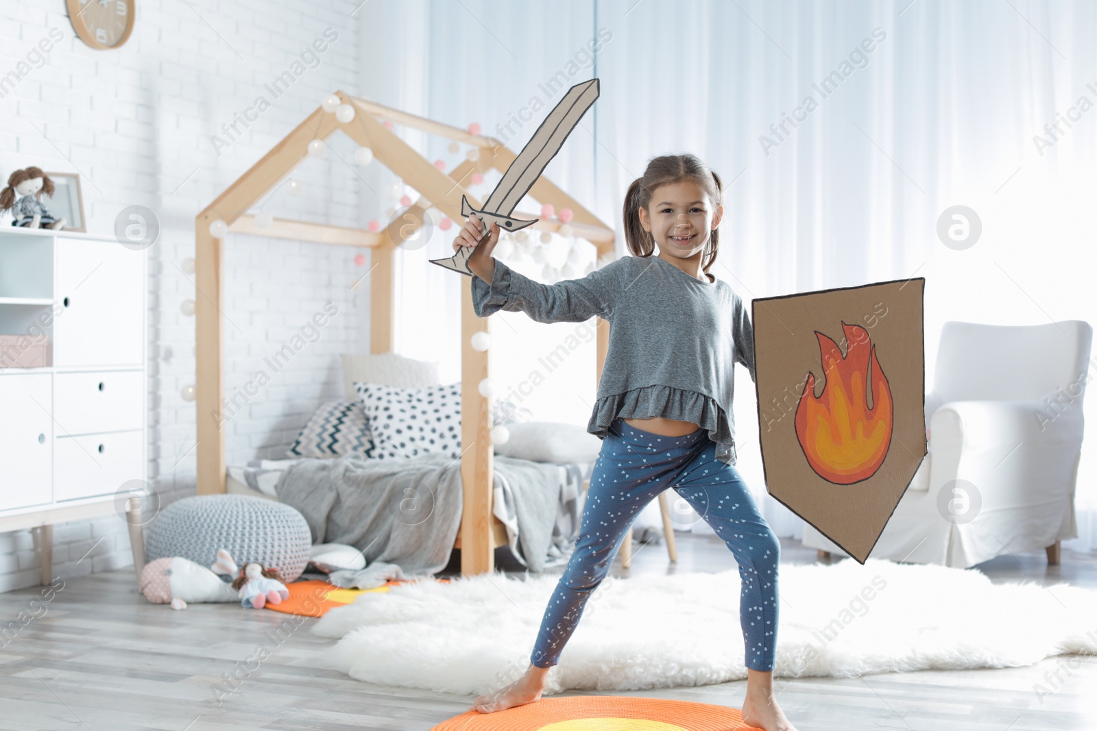 Photo of Cute little girl playing with cardboard armor in bedroom