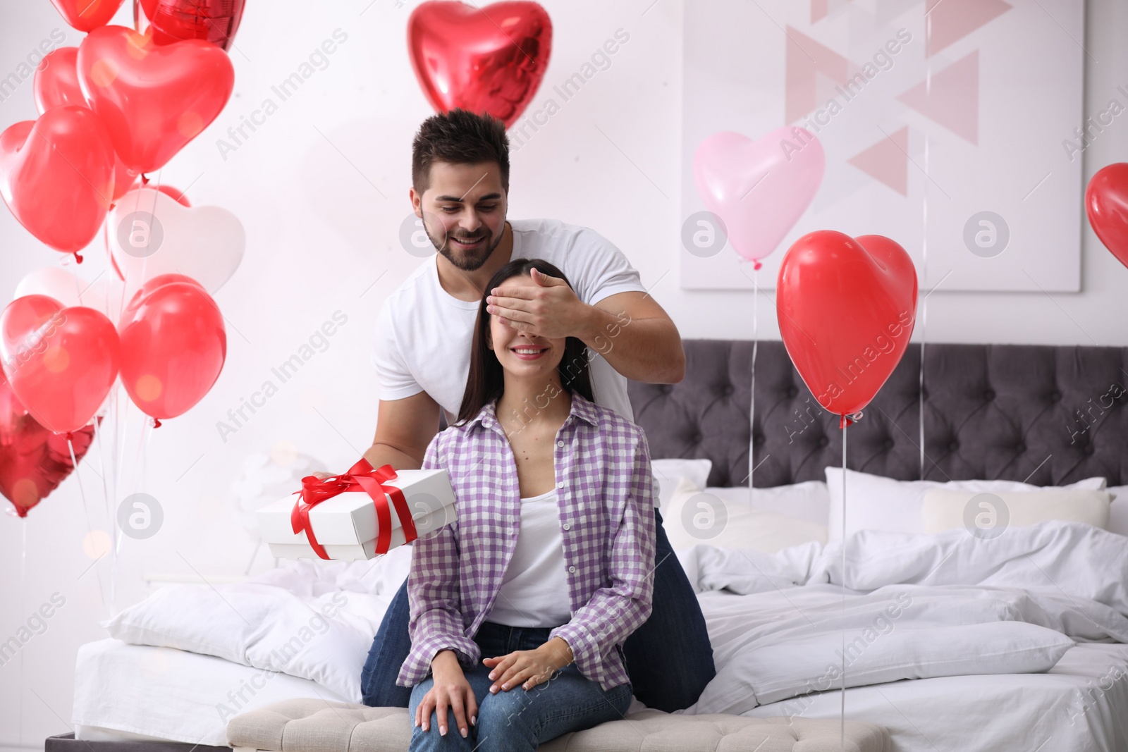 Photo of Young man presenting gift to his girlfriend in bedroom decorated with heart shaped balloons. Valentine's day celebration