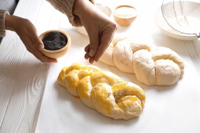Photo of Woman adding poppy seeds onto braided dough at white wooden table in kitchen, closeup. Cooking traditional Shabbat challah