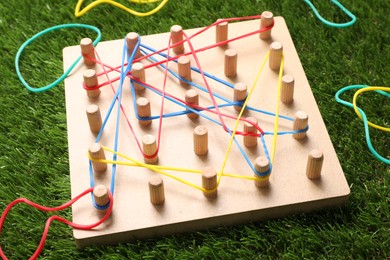 Photo of Wooden geoboard with rubber bands on artificial grass, closeup. Educational toy for motor skills development