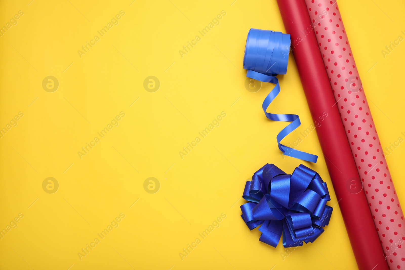 Photo of Rolls of colorful wrapping paper, ribbon and bow on yellow background, flat lay. Space for text