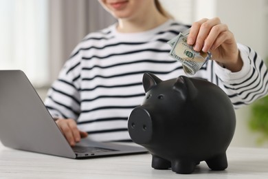Photo of Financial savings. Woman putting dollar banknote into piggy bank while using laptop at white wooden table indoors, closeup