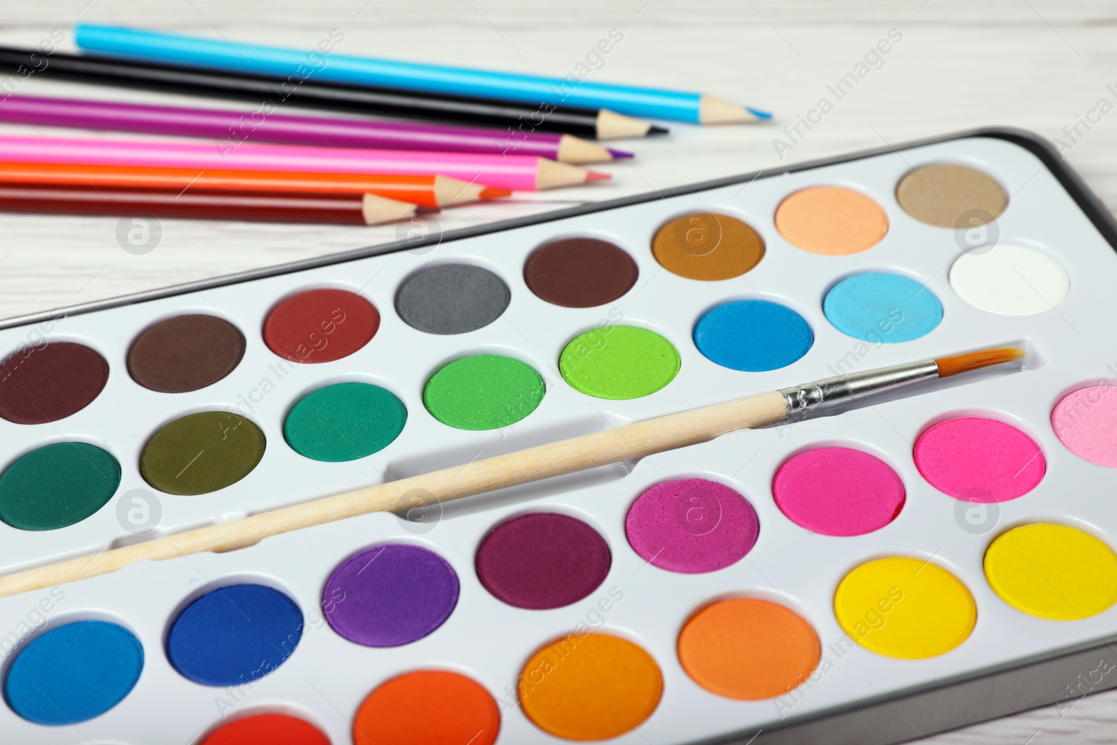 Photo of Watercolor palette with brush and colorful pencils on white wooden table, closeup