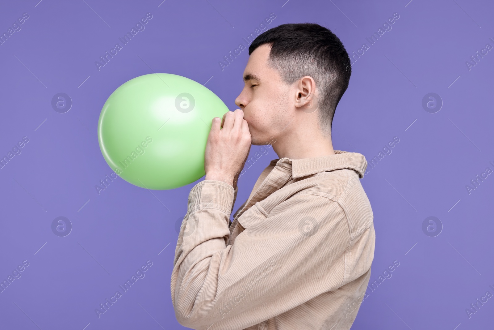 Photo of Young man inflating light green balloon on purple background