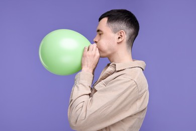Young man inflating light green balloon on purple background