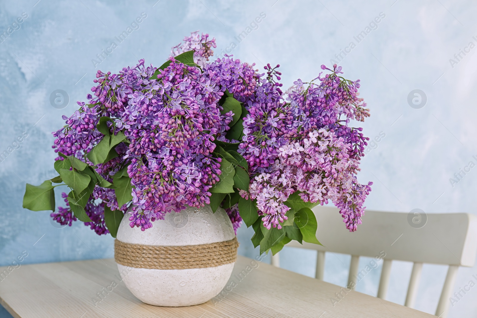 Photo of Vase with beautiful blossoming lilac on table against color background. Spring flowers