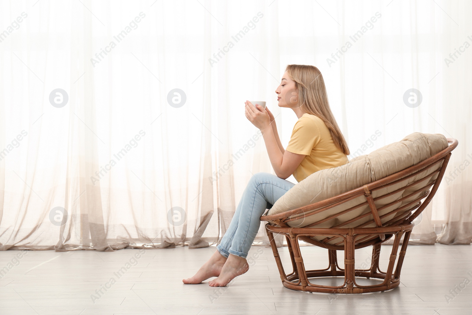 Photo of Beautiful young woman with cup of hot drink relaxing in papasan chair at home, space for text