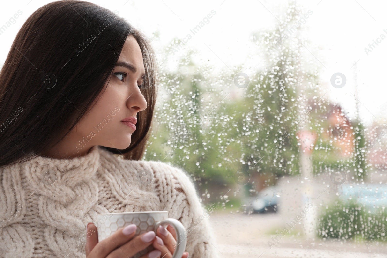 Photo of Sad beautiful woman with cup near window indoors on rainy day