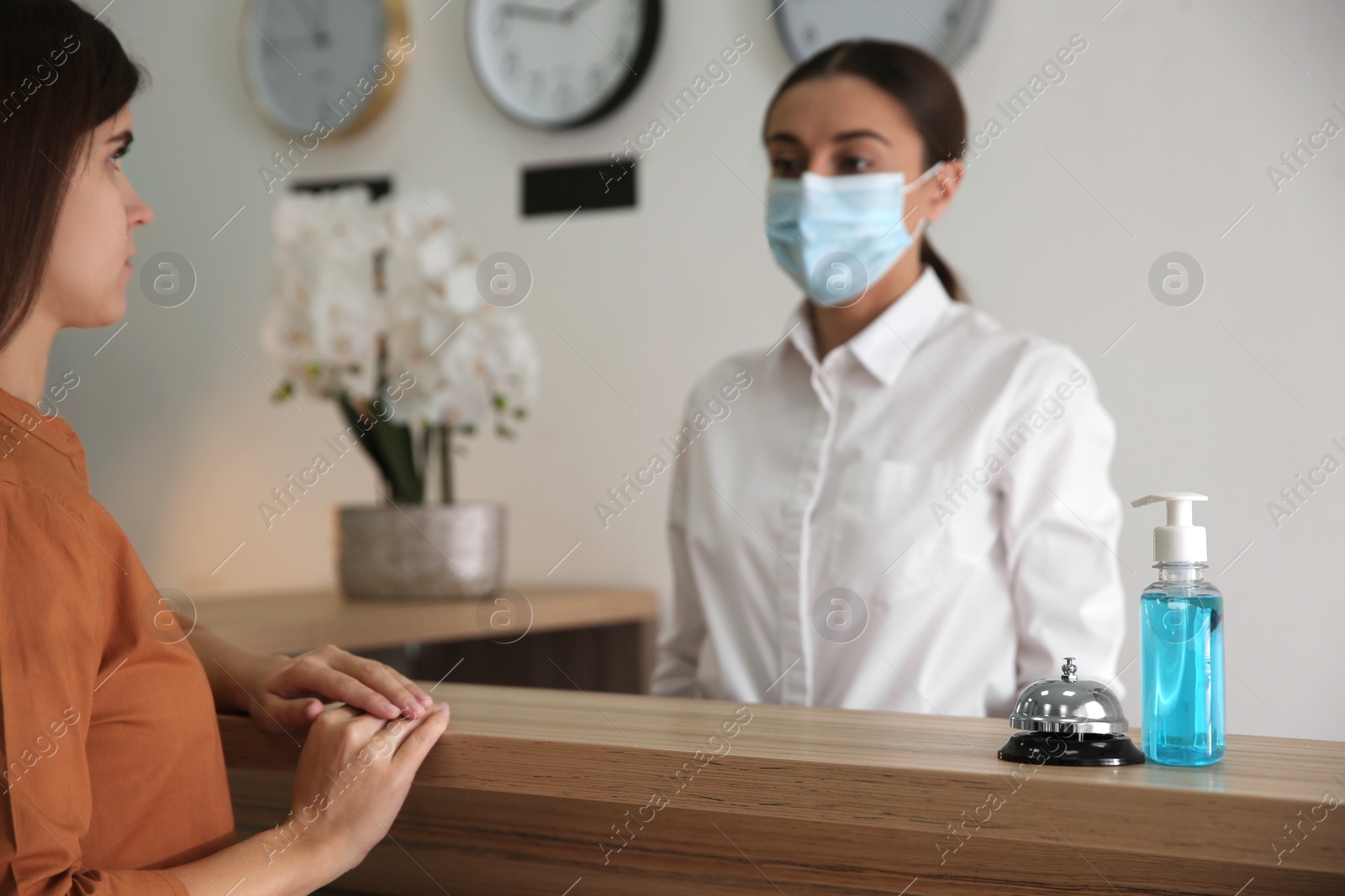 Photo of Receptionist with client at countertop in hotel, focus on dispenser bottle of antiseptic gel and service bell