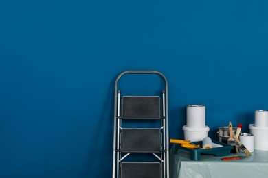 Photo of Metallic folding ladder and painting tools on table near blue wall, space for text